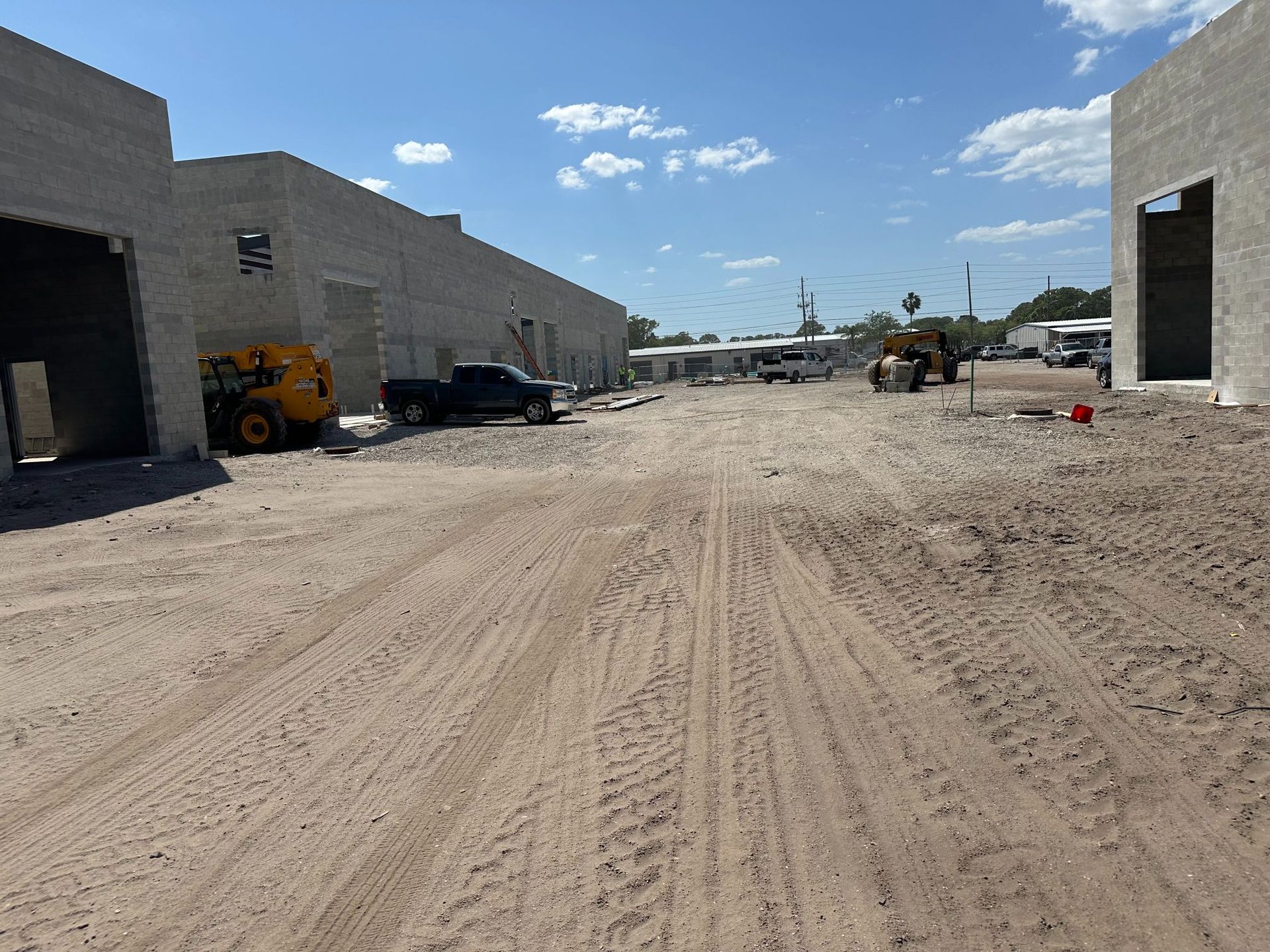 Construction site: dirt road between unfinished concrete buildings, vehicles, blue sky.