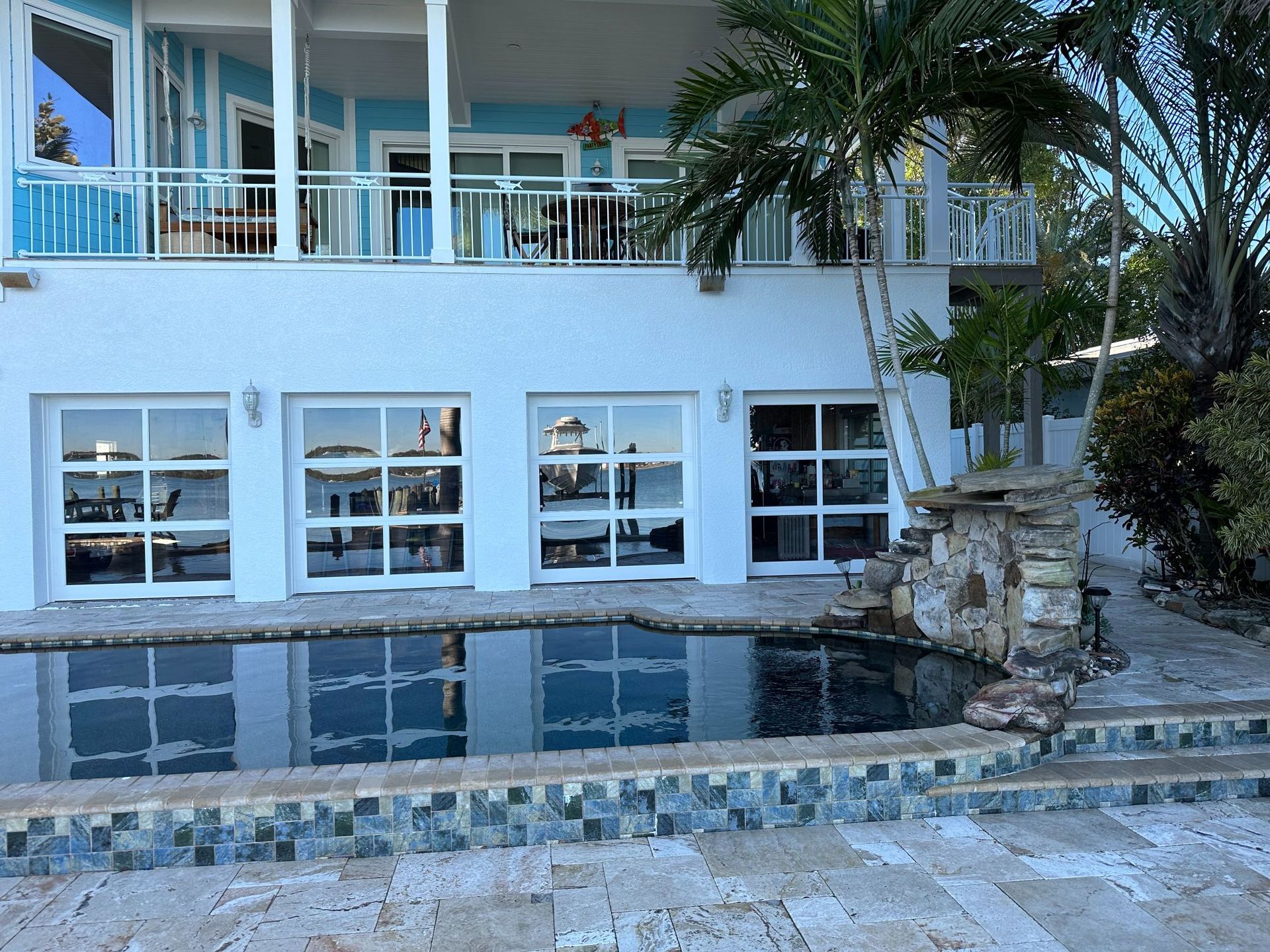 Two-story blue and white house with a pool. Large windows reflect the water. Palm trees and foliage surround.