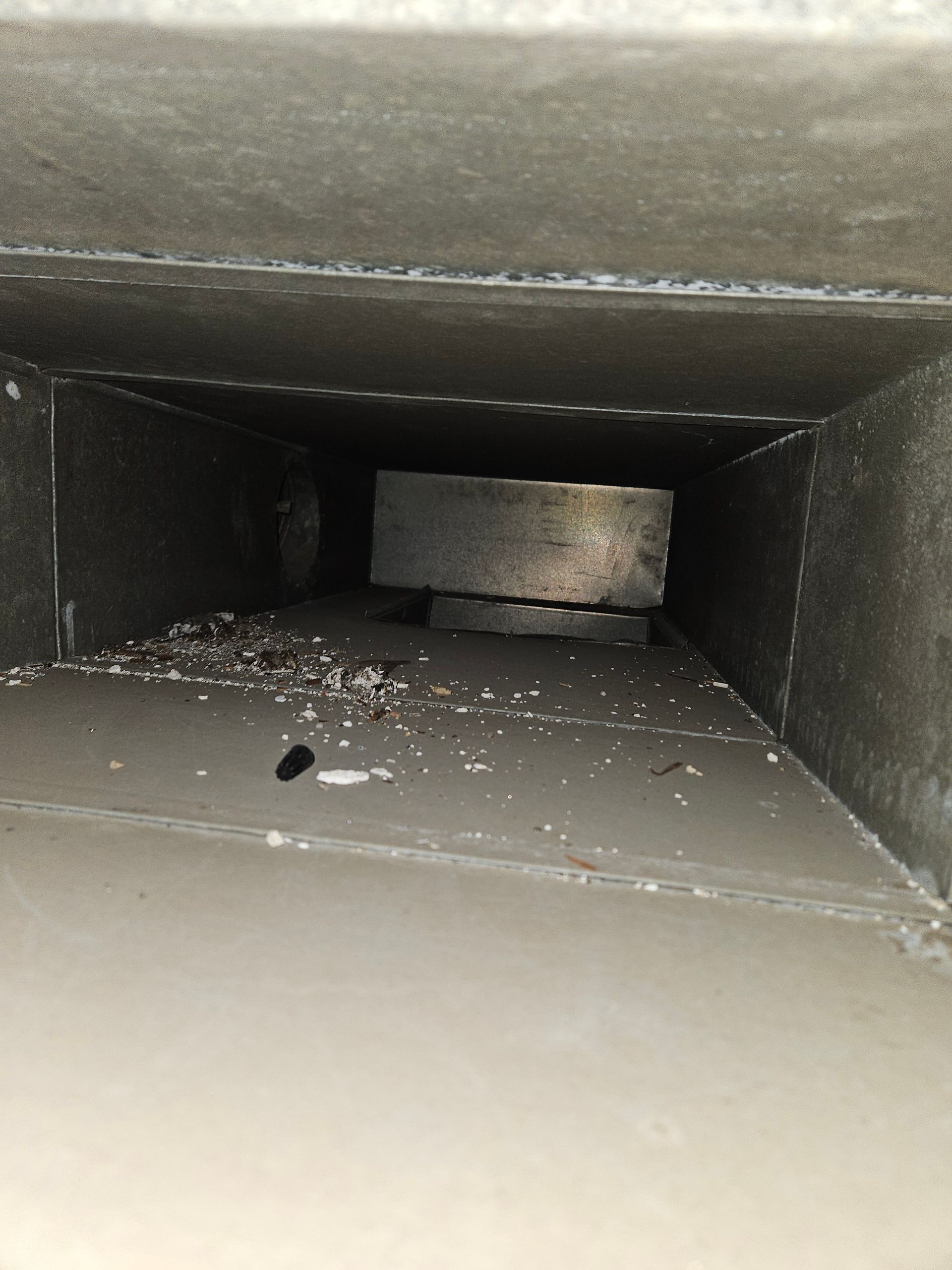 Inside view of a rectangular HVAC duct, showing dust and debris, with a metal component partially blocking the airflow.