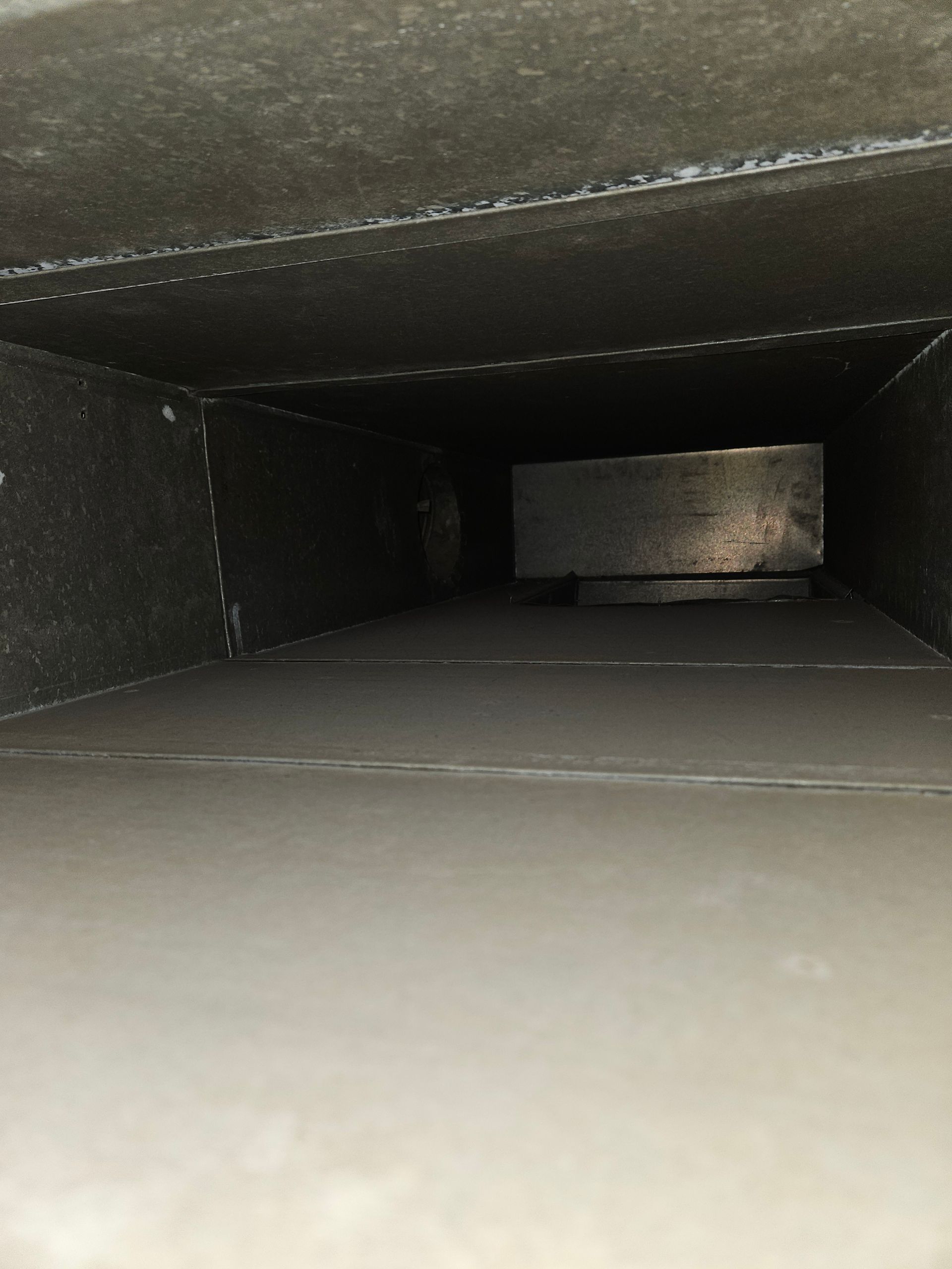 Inside view of a dark rectangular duct, showing a metal component and dust along the sides.