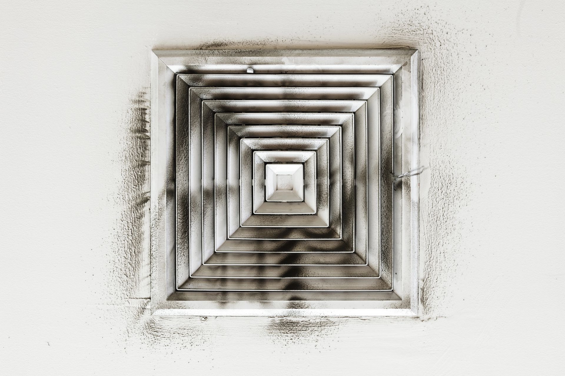 Dirty air vent in a white ceiling. Black and gray grime covers the vent and surrounding surface.