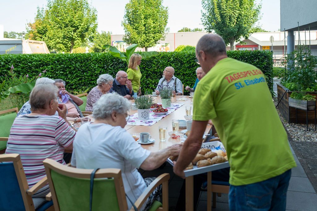 Gäste der Tagespflege St. Elisabeth Bühl sitzen zum Nachmittagskaffee im Garten und werden von Betreuungskräften bedient.