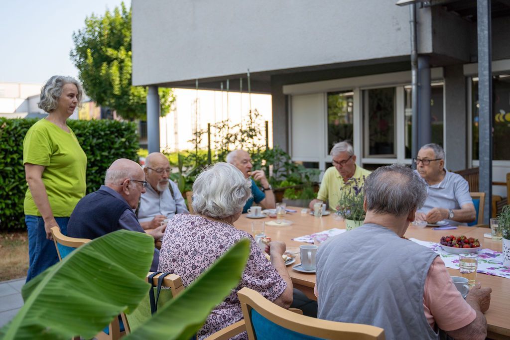 Gäste der Tagespflege St. Elisabeth Bühl sitzen im Garten beim Nachmittagscafe und eine Betreuungskraft steht daneben.