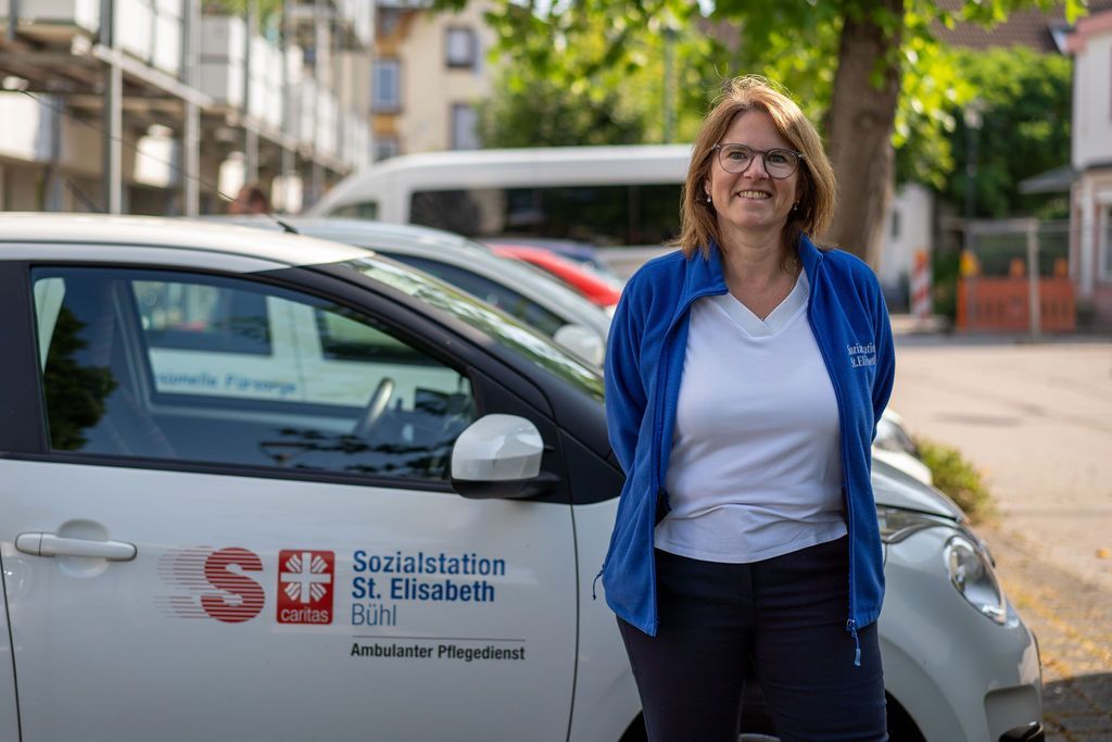 Pflegefachkraft steht neben Dienstwagen von Essen auf Rädern.