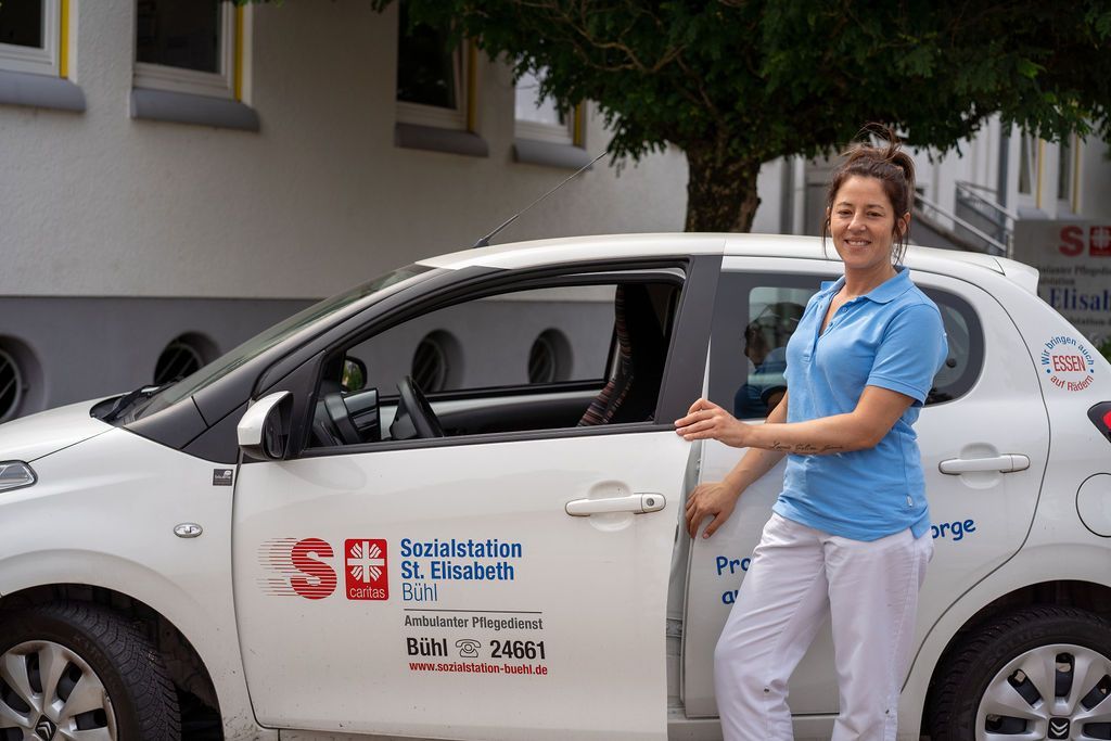 Pflegefachkraft steht in Arbeitskleidung neben Dienstwagen.