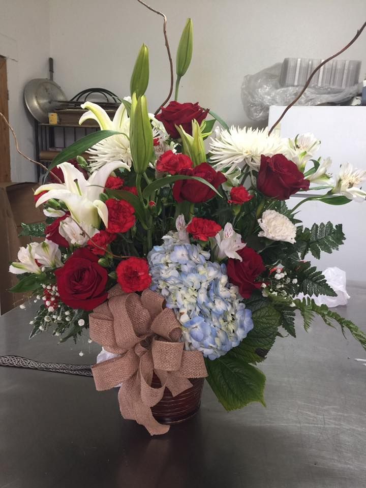Flower arrangement in a basket with red roses, white lilies, blue hydrangea, and a burlap bow.