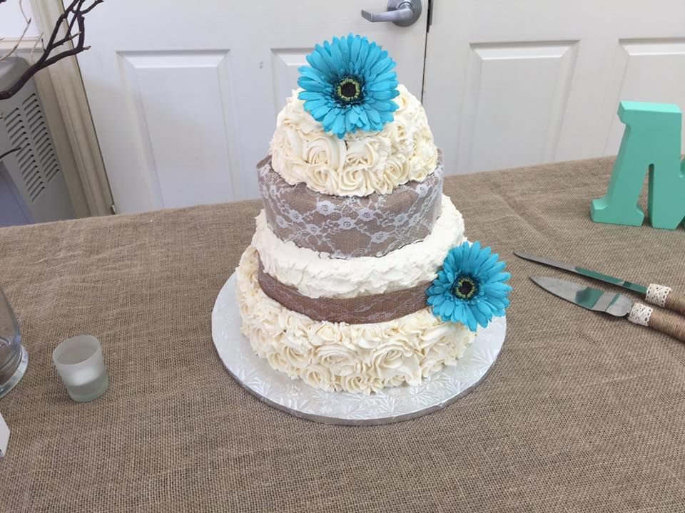 Three-tiered cake with cream frosting, brown ribbon, and blue flower decorations on a burlap-covered table.