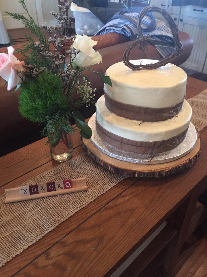 Two-tiered cake with brown bands on a wooden slab, decorated with a branch heart. Flowers and game tiles on a table.