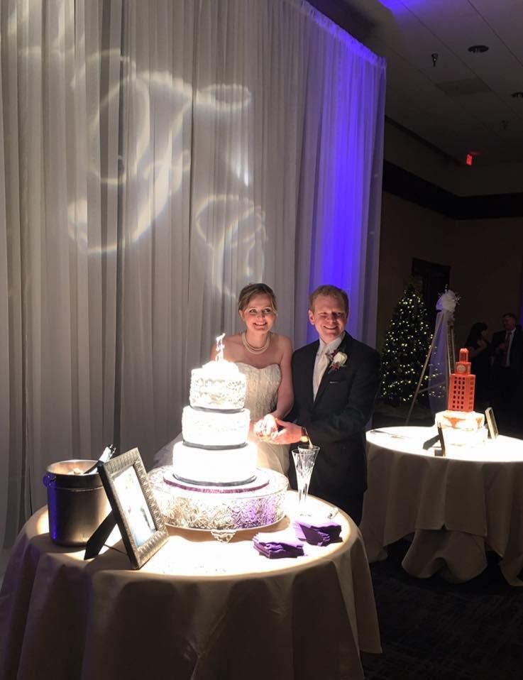 Bride and groom cutting wedding cake at reception, purple and white decorations.