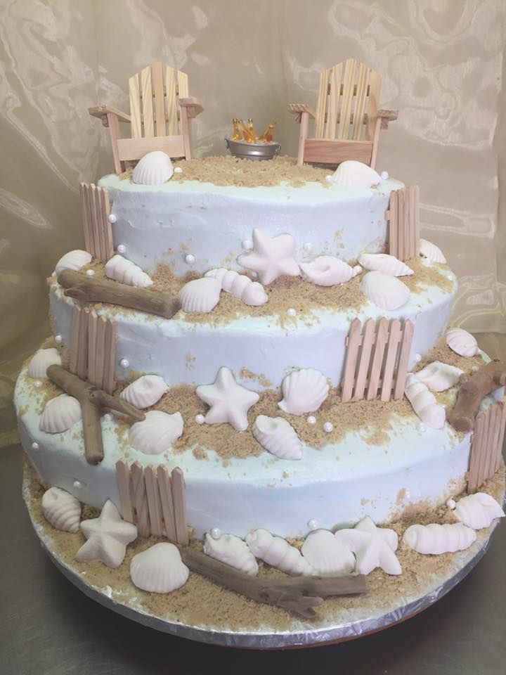 Three-tiered beach-themed cake with light blue frosting, seashells, sand, small wooden fences, and Adirondack chairs.