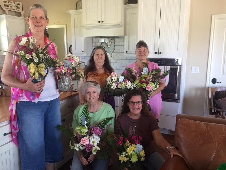 Six people holding floral bouquets in a kitchen setting. They smile, with white cabinets and a brick backsplash visible.