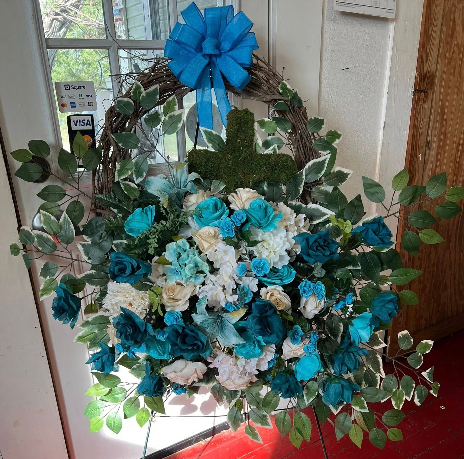 Wreath with blue bow and teal and white flowers, cross, and greenery on door.