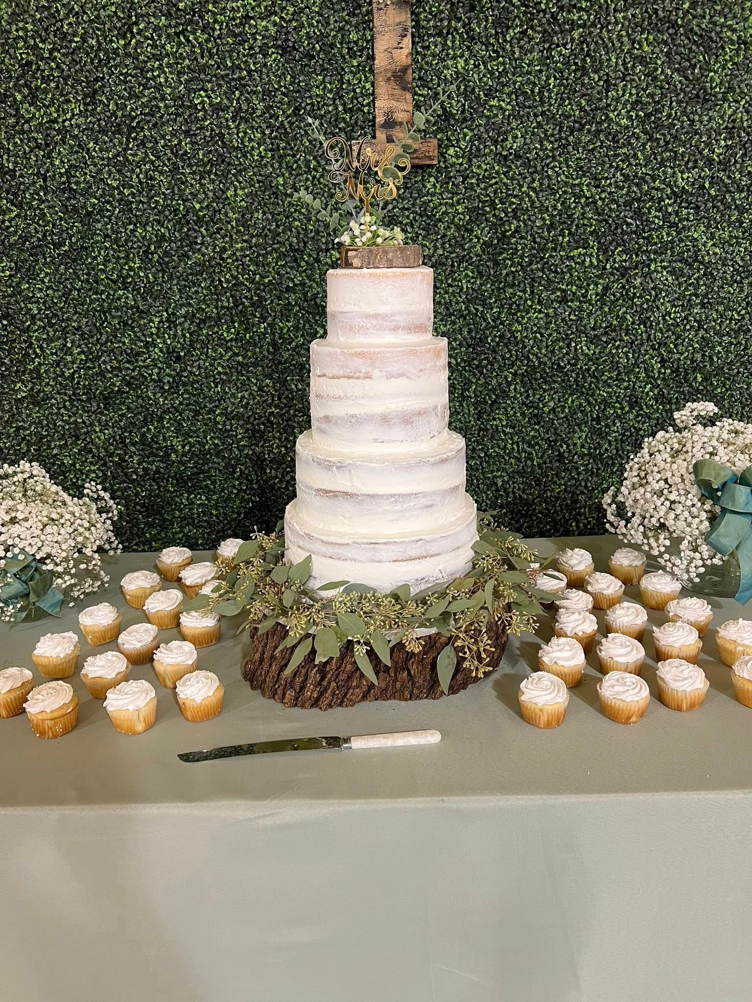 Naked-style wedding cake on a wooden base with cupcakes. Greenery and flowers decorate the table.