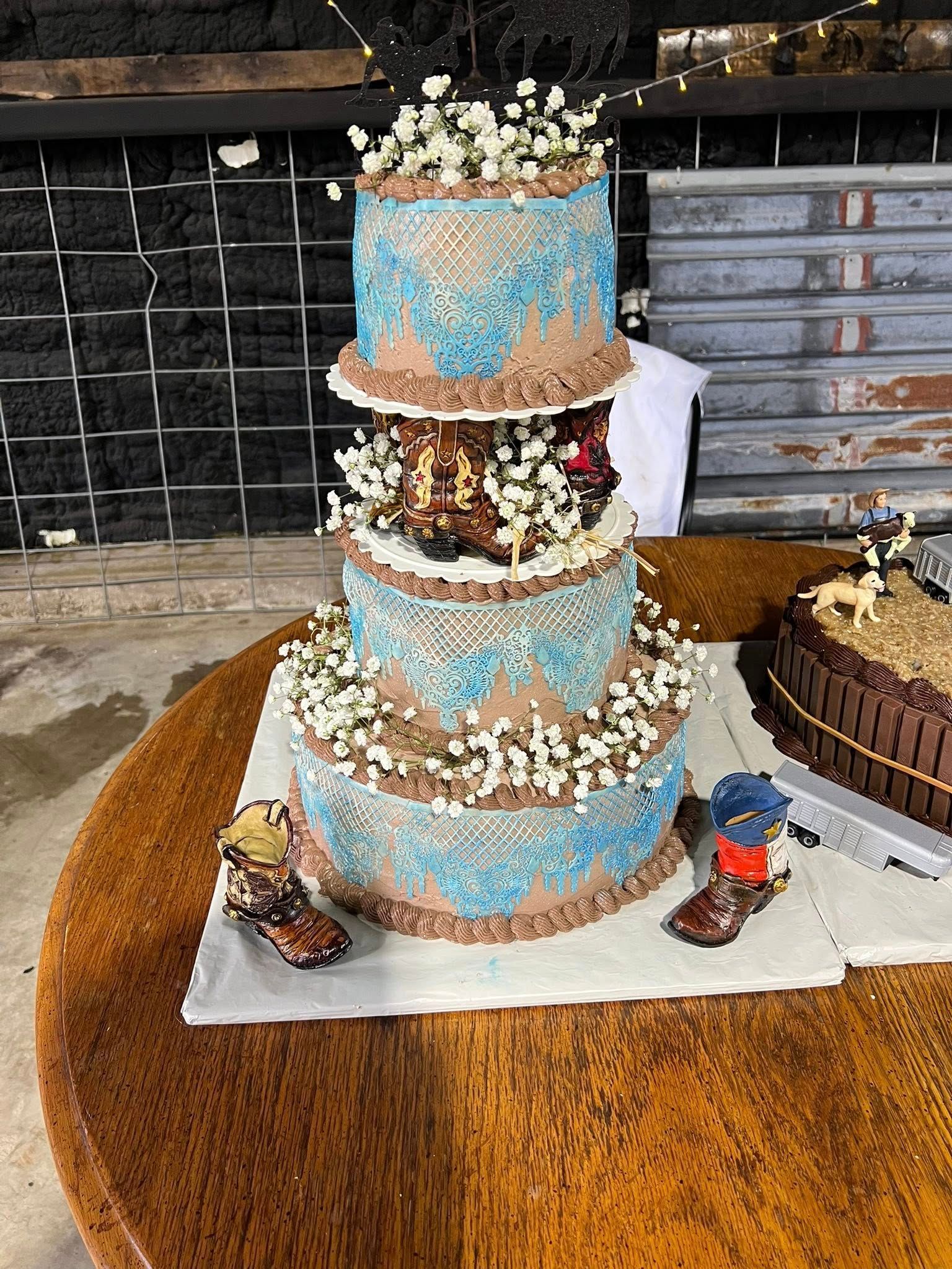 Four-tiered cake with blue lace design, adorned with white flowers. Cowboy boot figures sit at the base.