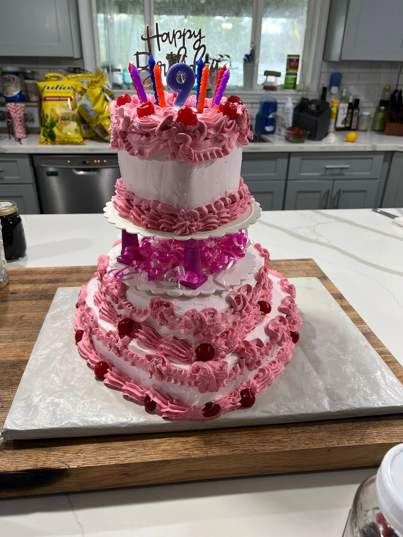 Three-tiered pink birthday cake with candles and cherries on a wooden board in a kitchen.