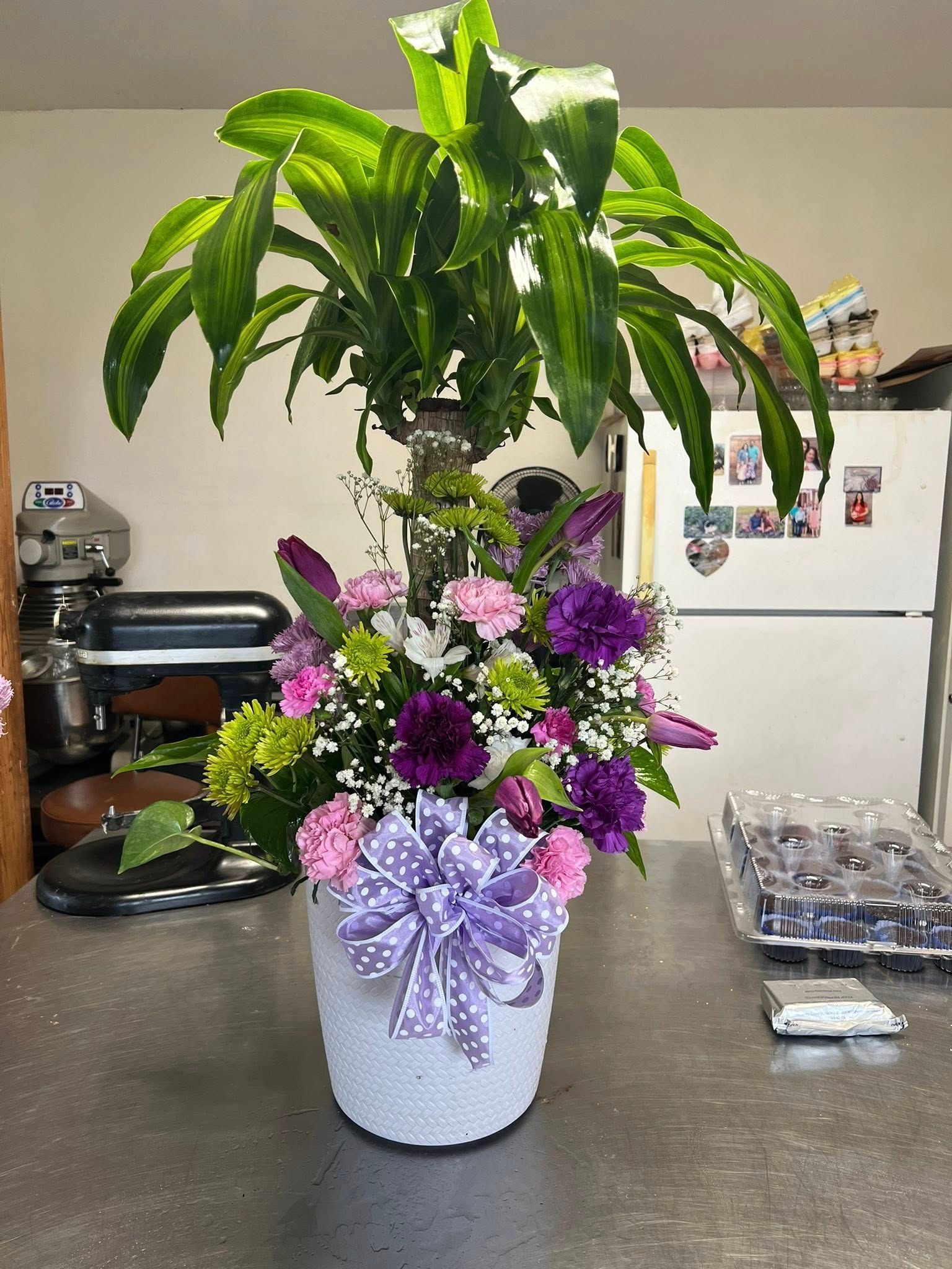 Floral arrangement in a white, textured pot with a purple bow. Includes green leaves, purple and pink flowers.