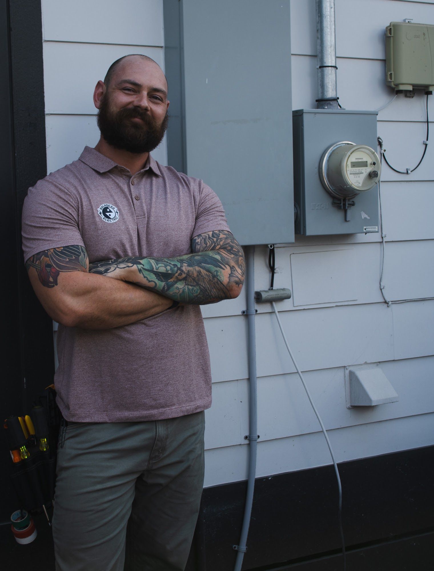 Man with arm tattoos and beard stands near electrical panel on a white building exterior.