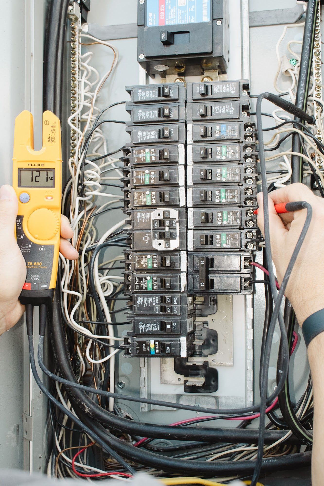 Person using a multimeter to test a circuit breaker in an electrical panel. Wires and breakers are visible.