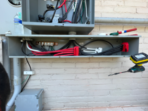 Electrical wiring project outdoors; open metal box, exposed wires, tools, and water bottle on a brick wall.