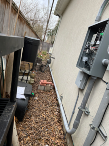 Exterior electrical panel on a beige wall, next to a narrow path with debris and a wooden fence.