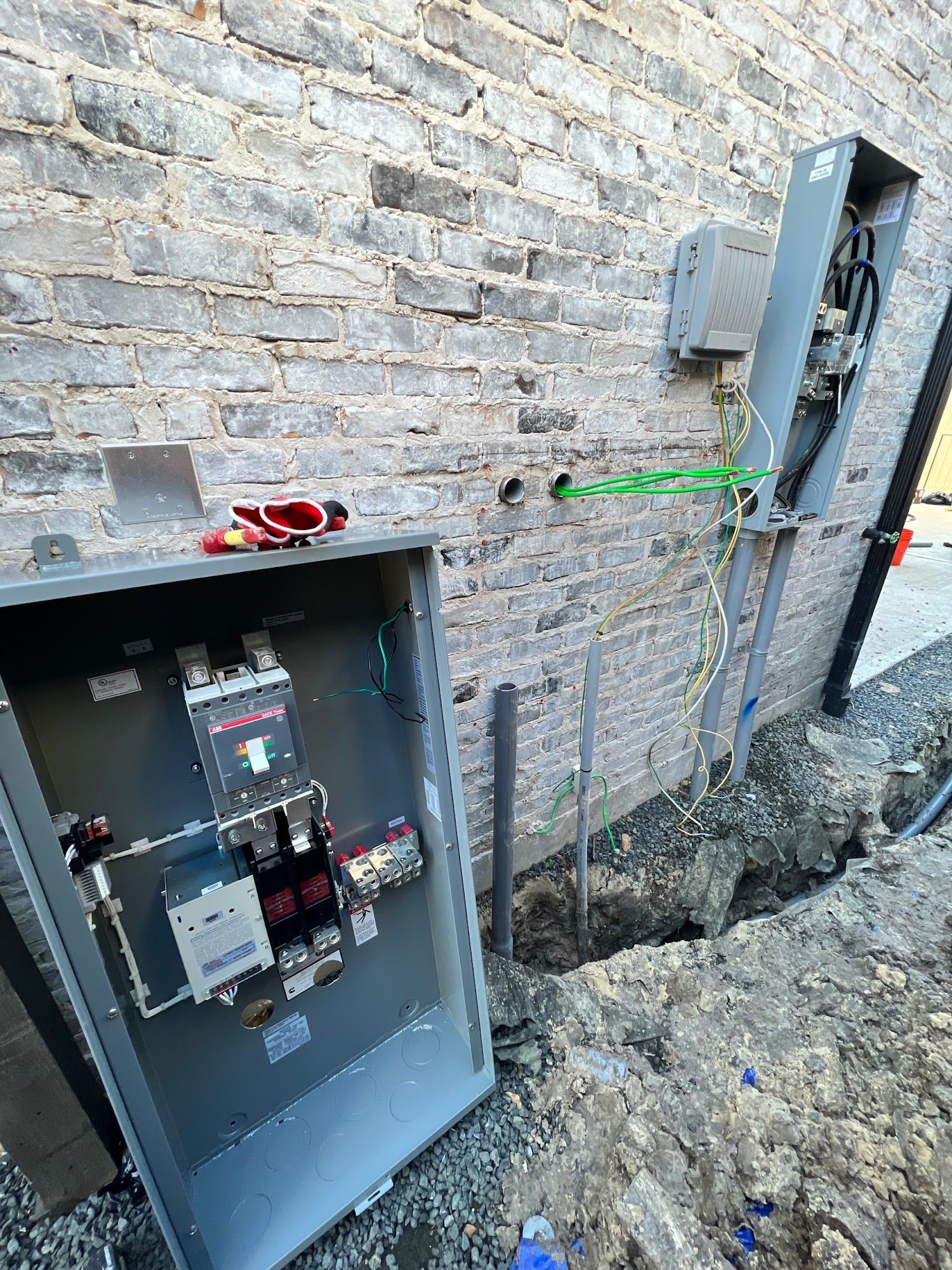 Electrical equipment installation: open metal box exposing circuit breakers and wiring on brick wall. Trench visible.