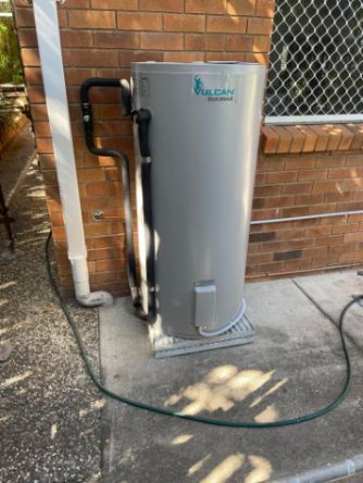 A Gray Rheem Hot Water Tank Against a Brick Wall, With Pipes and a Hose — Hales Plumbing & Roofing In The Range, QLD