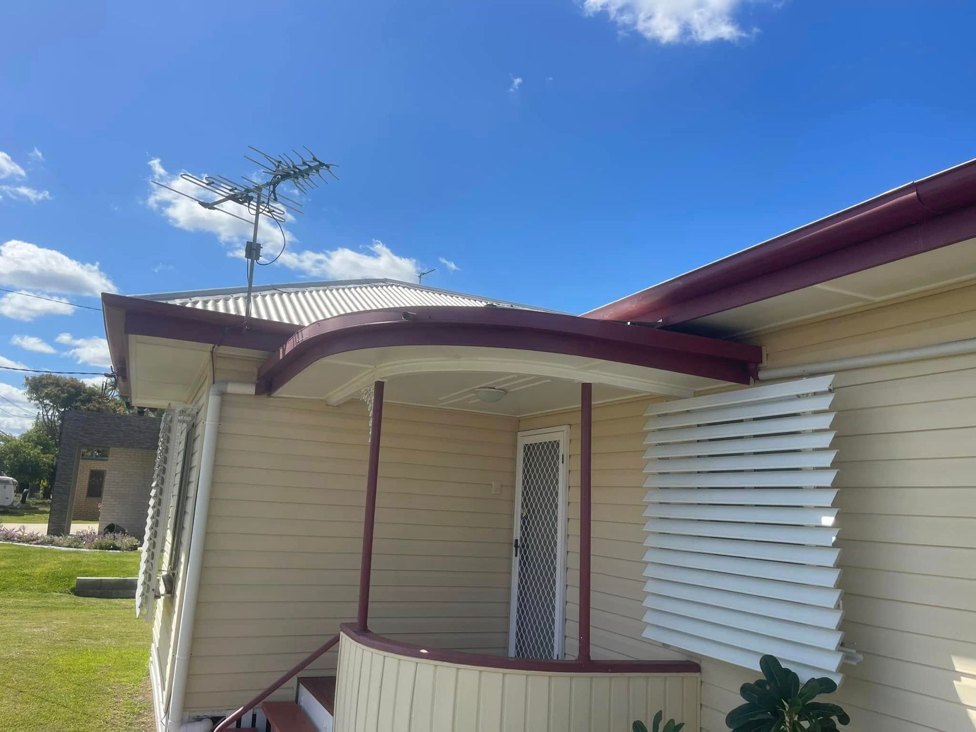 A light yellow house with a rounded porch, maroon trim, a white louvered window shutter, and a TV antenna on the roof — Hales Plumbing & Roofing In Gracemere, QLD