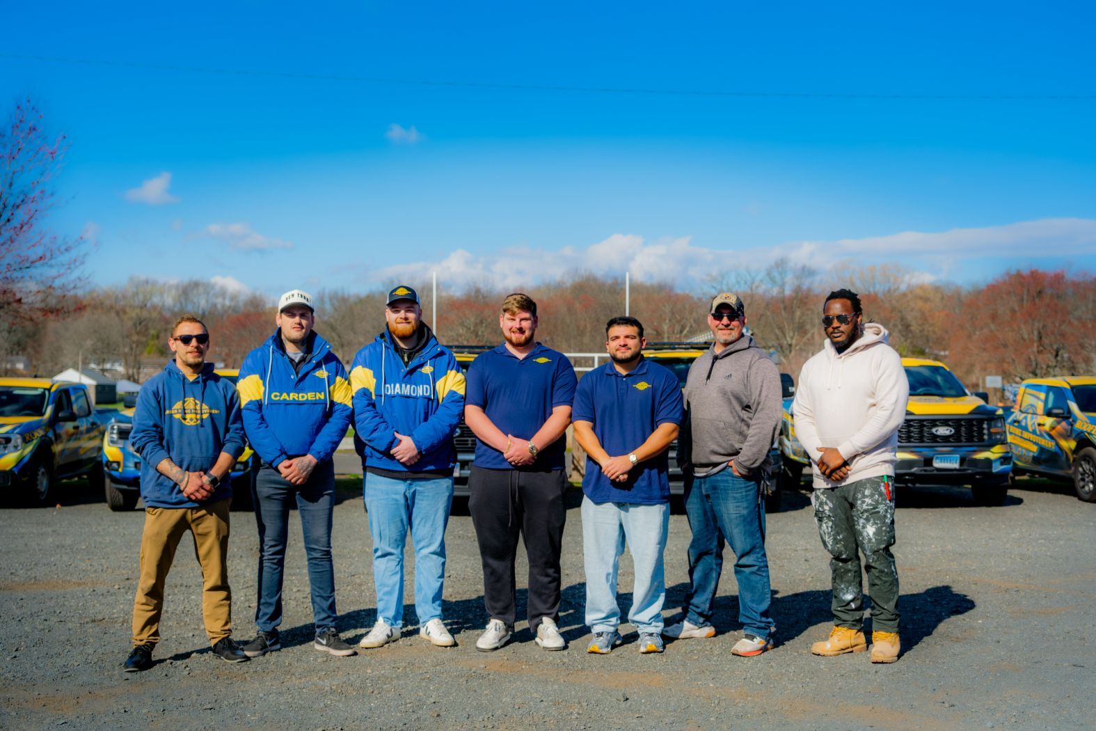 Seven people standing in a parking lot with blue-and-yellow cars behind them under a bright sky