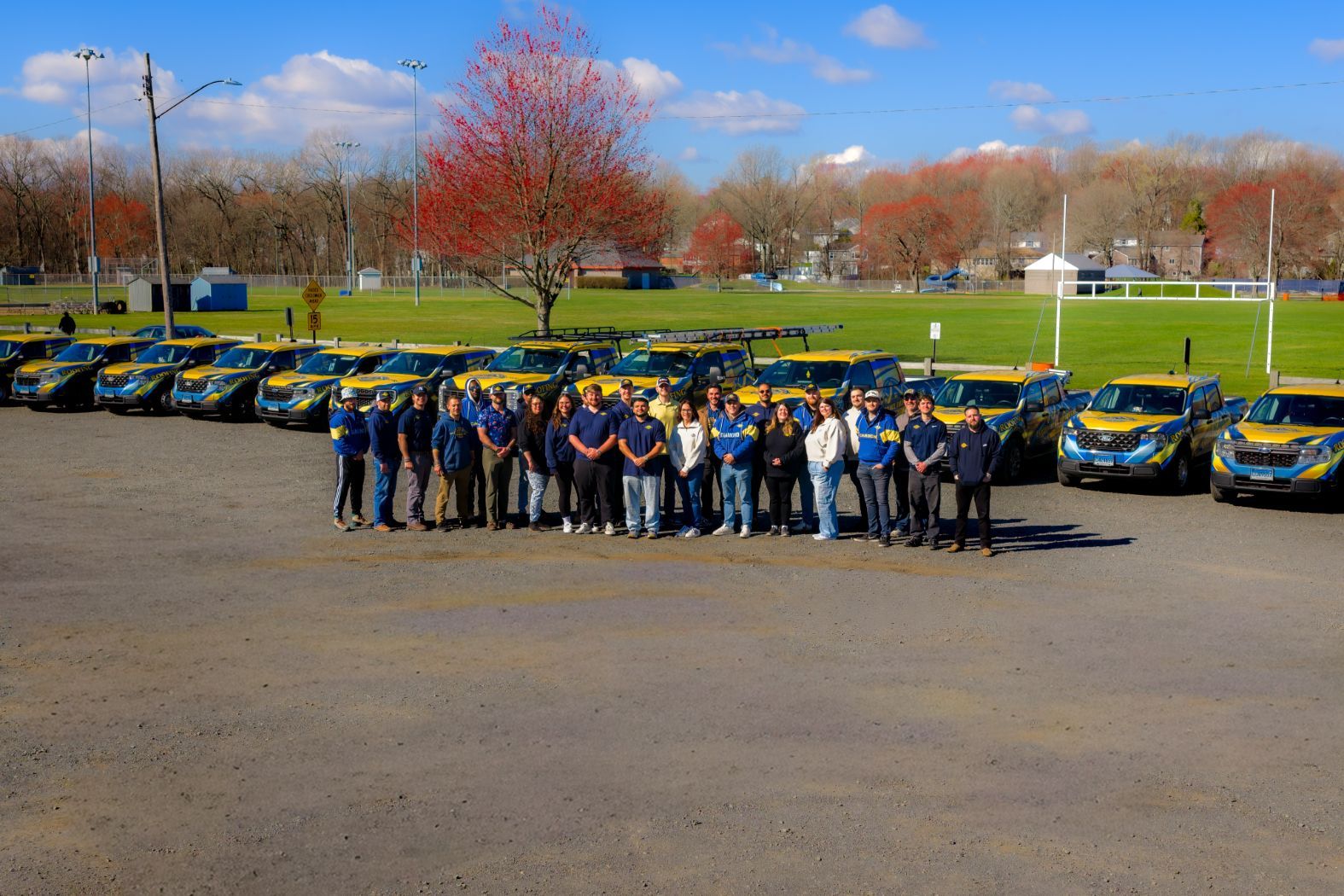 Group of people posing in front of yellow-and-blue cars in a parking lot under a blue sky.