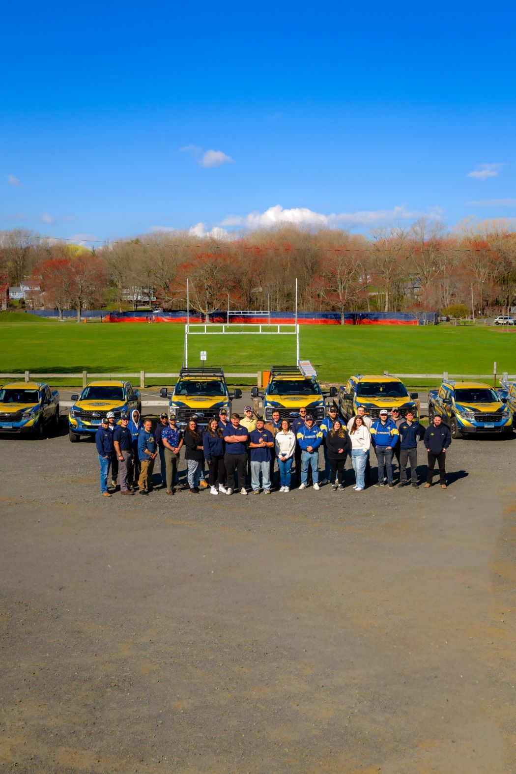 Group of people posing with black cars on a sunny field, with a track and trees in the background