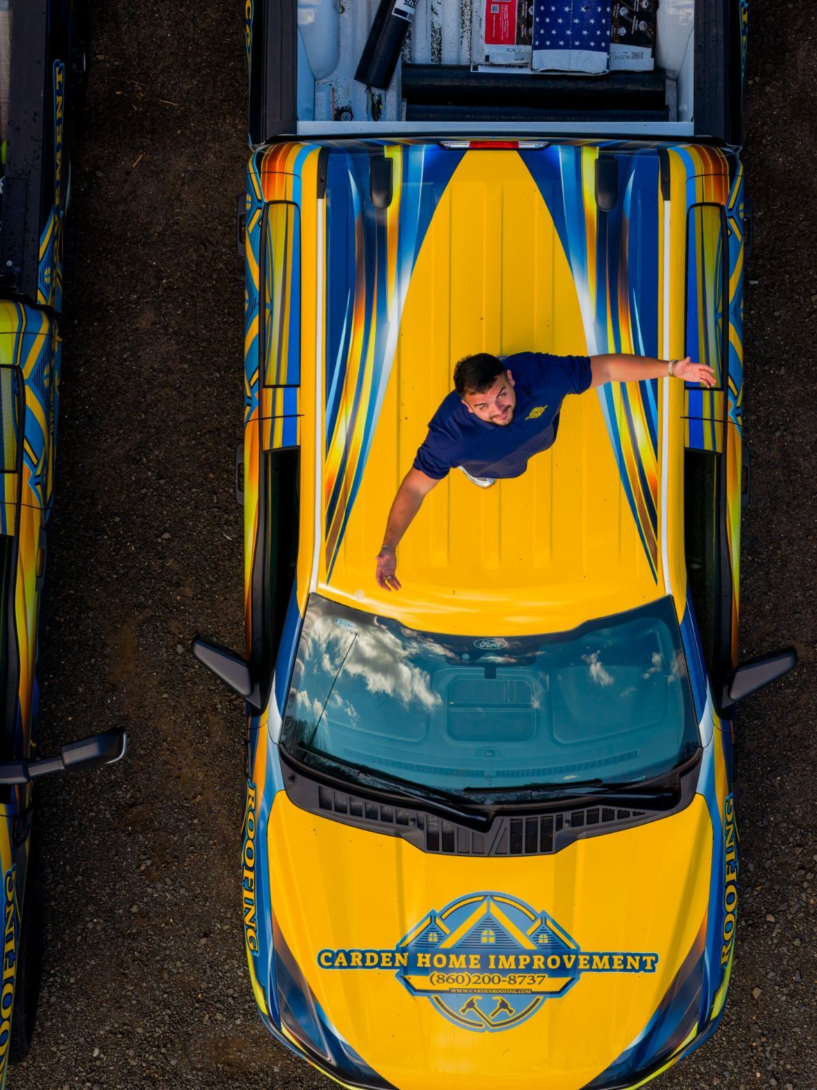 Person standing on a yellow-and-blue car roof beside a trailer, viewed from above.