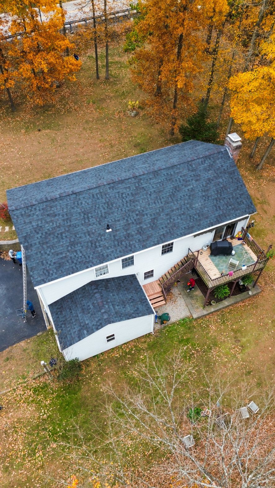 Aerial view of a white house with a dark roof beside autumn trees and a small deck.