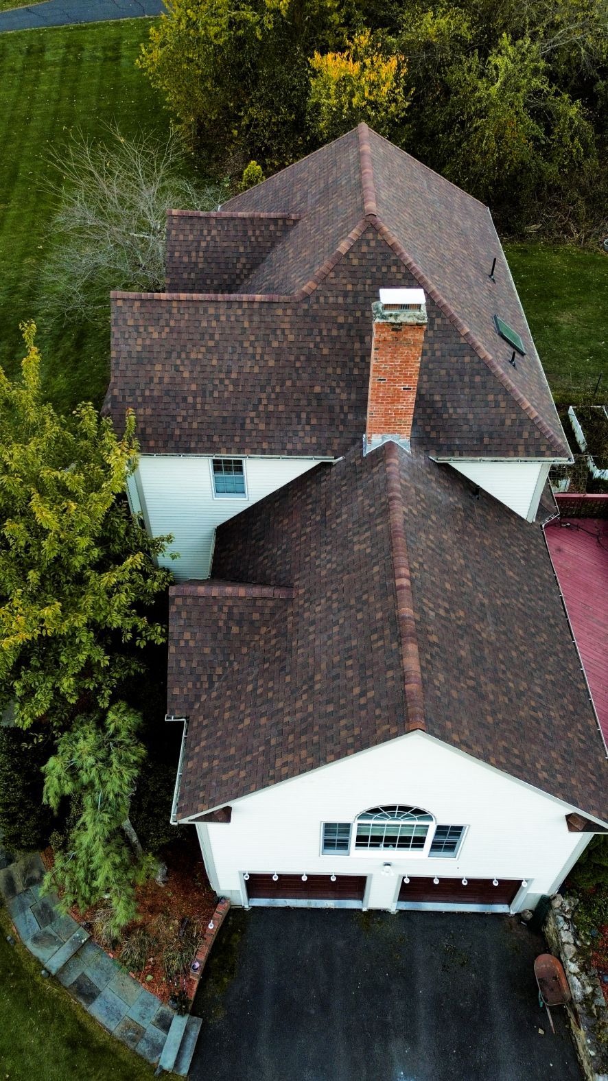 Aerial view of a white house with a brown shingled roof and driveway, surrounded by trees.