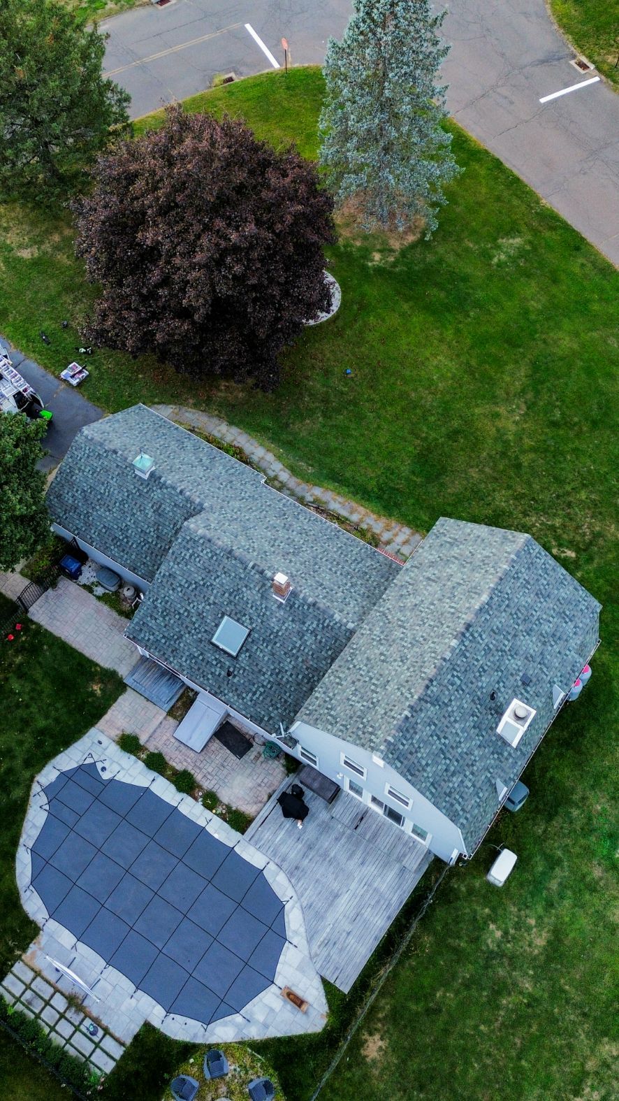 Aerial view of a house with a gray roof, solar panels, and a lawn with trees beside a driveway.