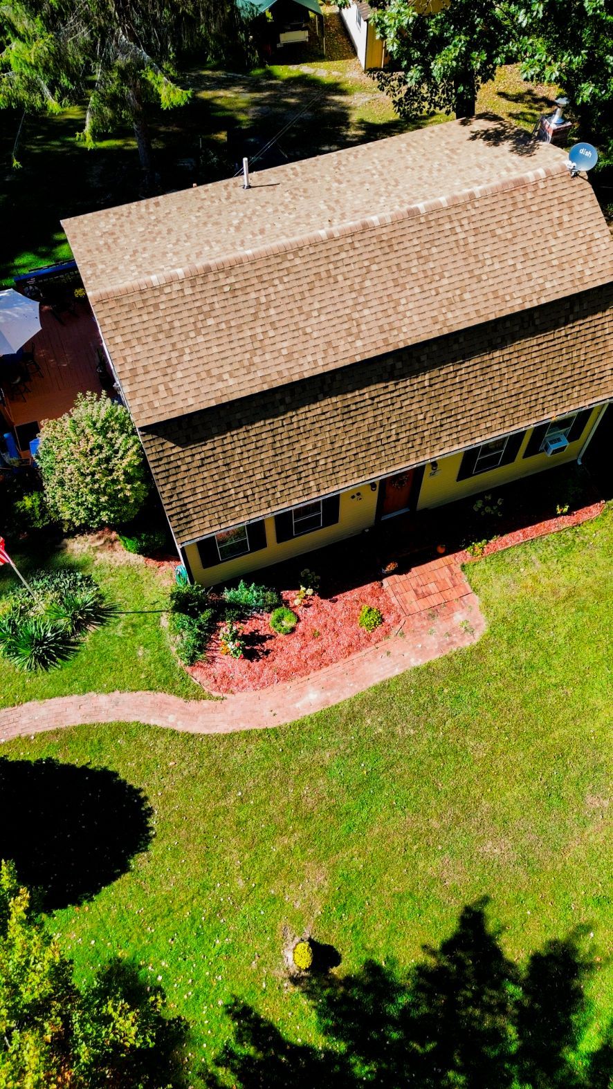 Aerial view of a tan-roofed house surrounded by green lawn, trees, and a small red brick patio.