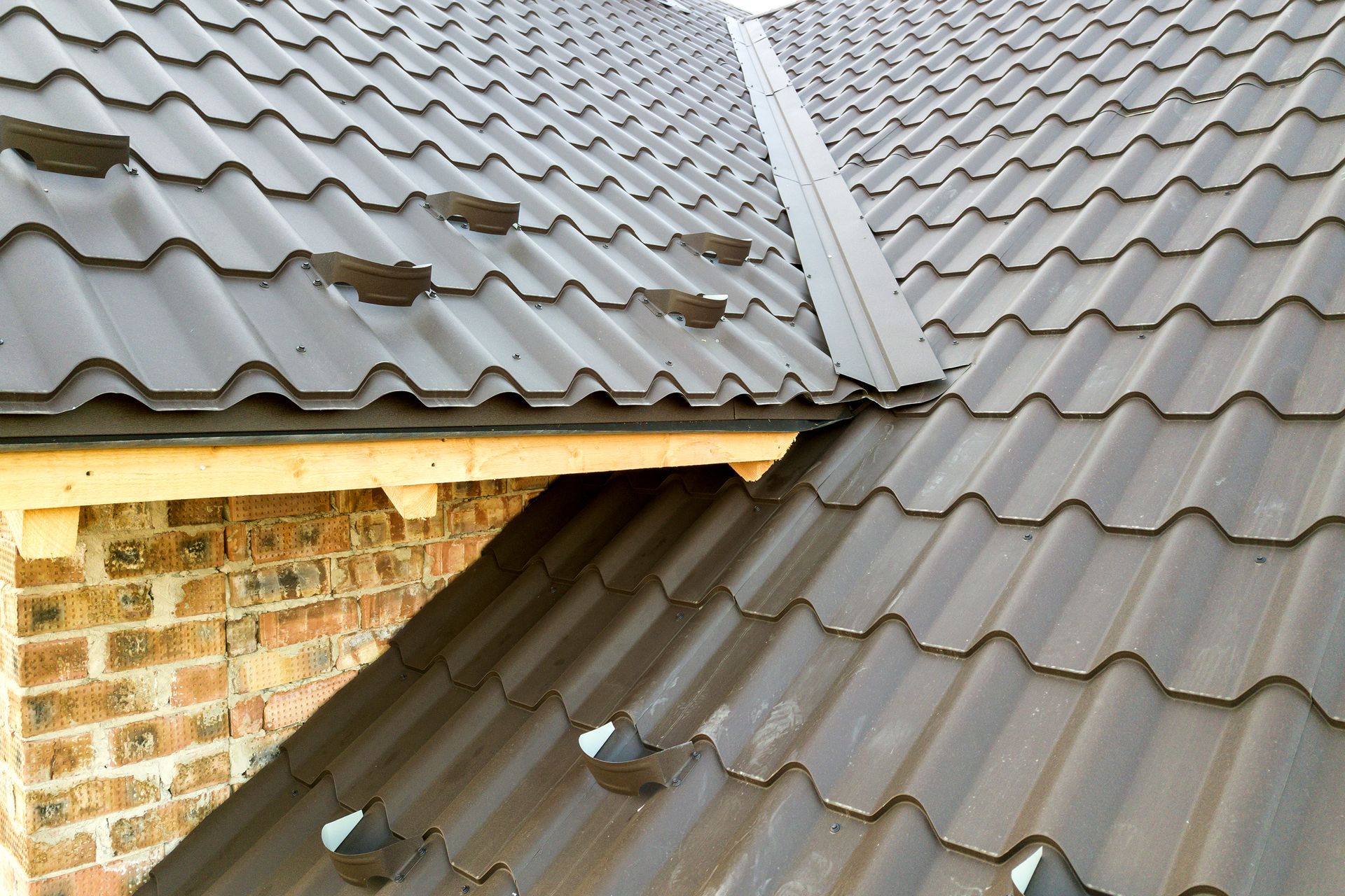 Two gray tiled roof sections meeting beside a brick wall, with a visible roof seam and flashing.