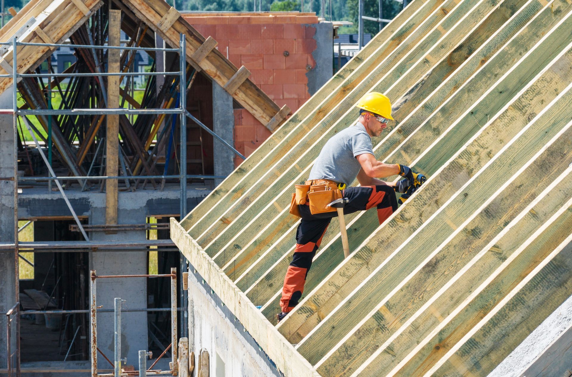 Construction worker on a roof frame with a nail gun, wearing a yellow hard hat and safety harness.