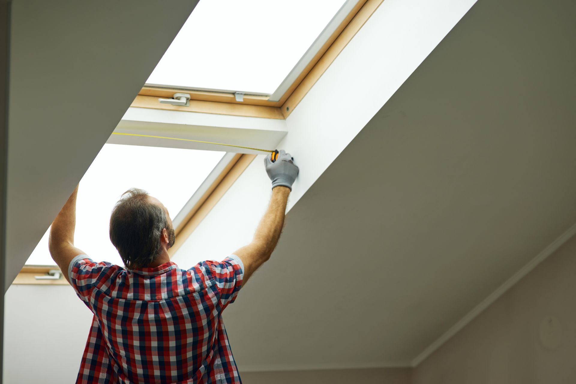 Person with a beard and plaid shirt working on a skylight, wearing gloves, indoors.