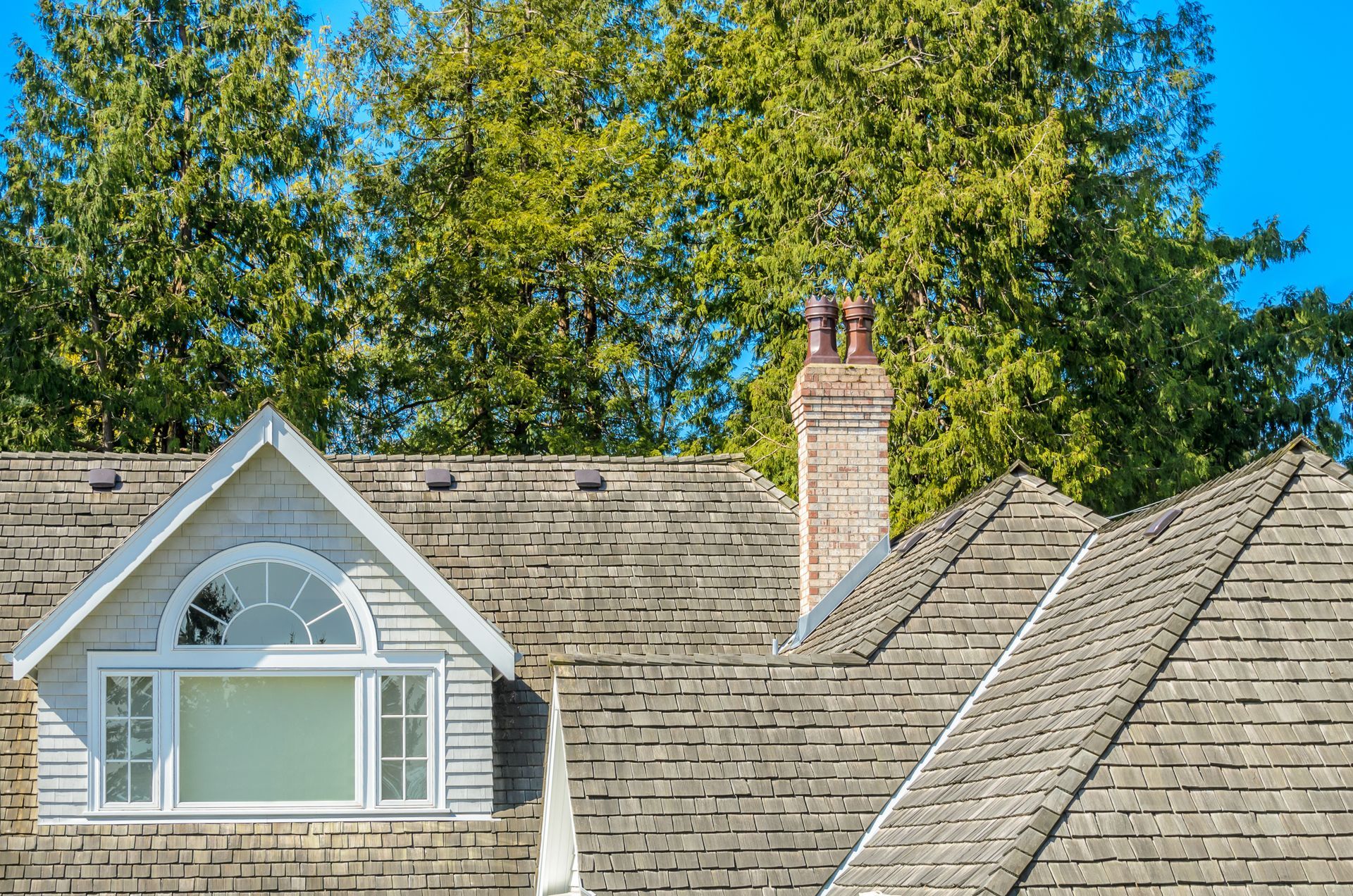 Gray shingled house roof with dormer window, brick chimney, and tall green trees under a blue sky