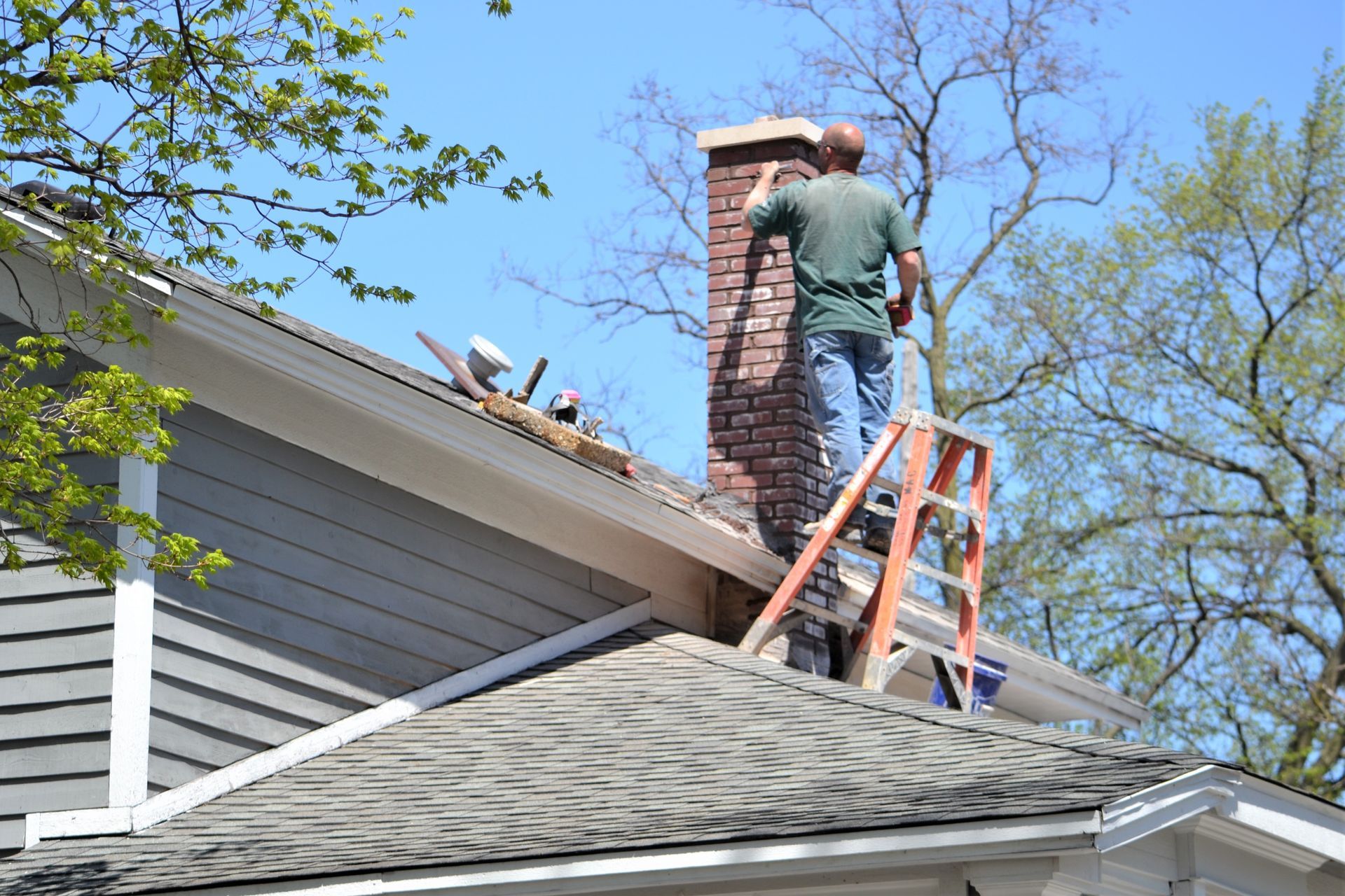 Person on ladder repairs chimney on a house roof. Blue sky, tools, and trees visible.