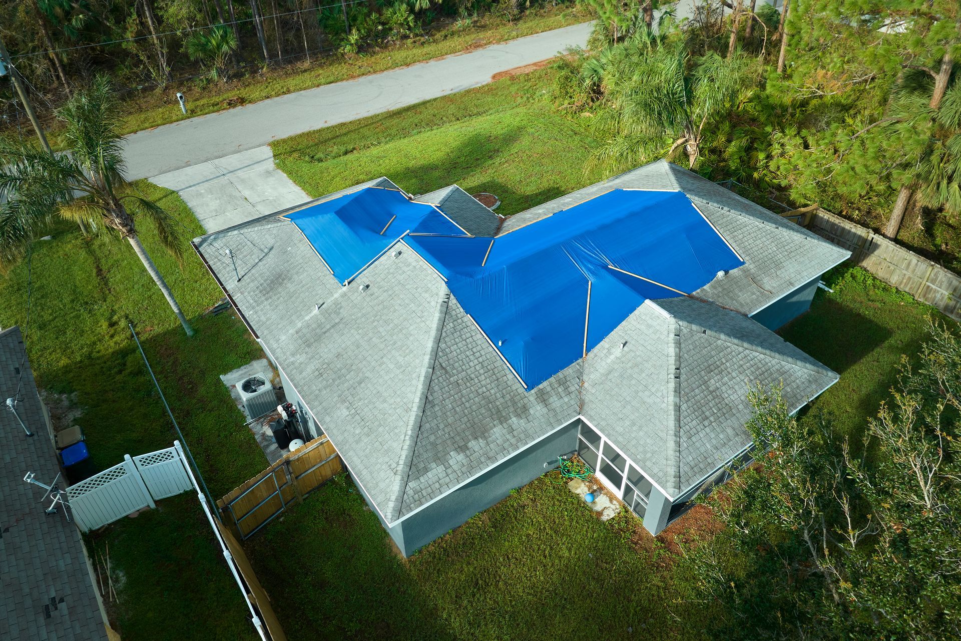 Aerial view of a house with a gray roof partially covered with blue tarps. A driveway and green yard are visible.