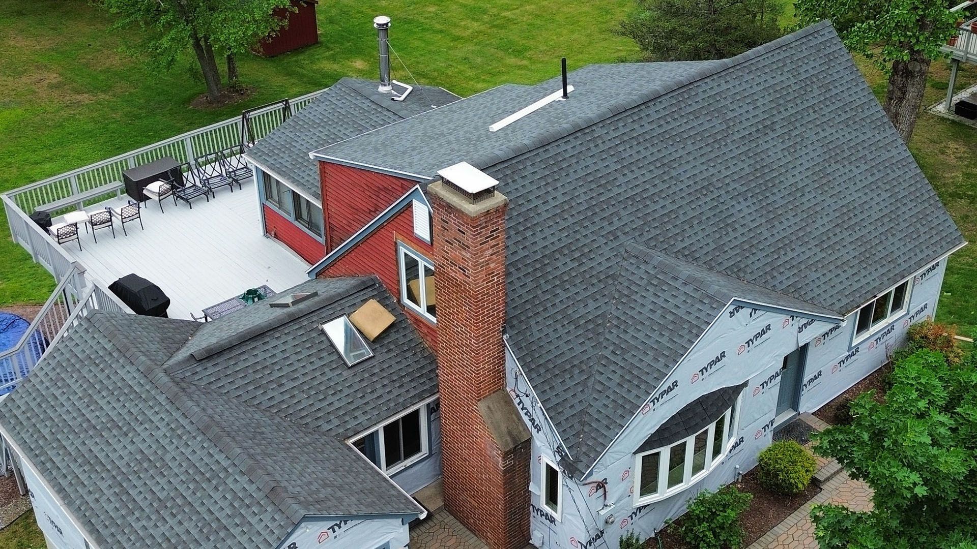 Aerial view of a large gray-roofed house with a red brick chimney and damaged roof section.