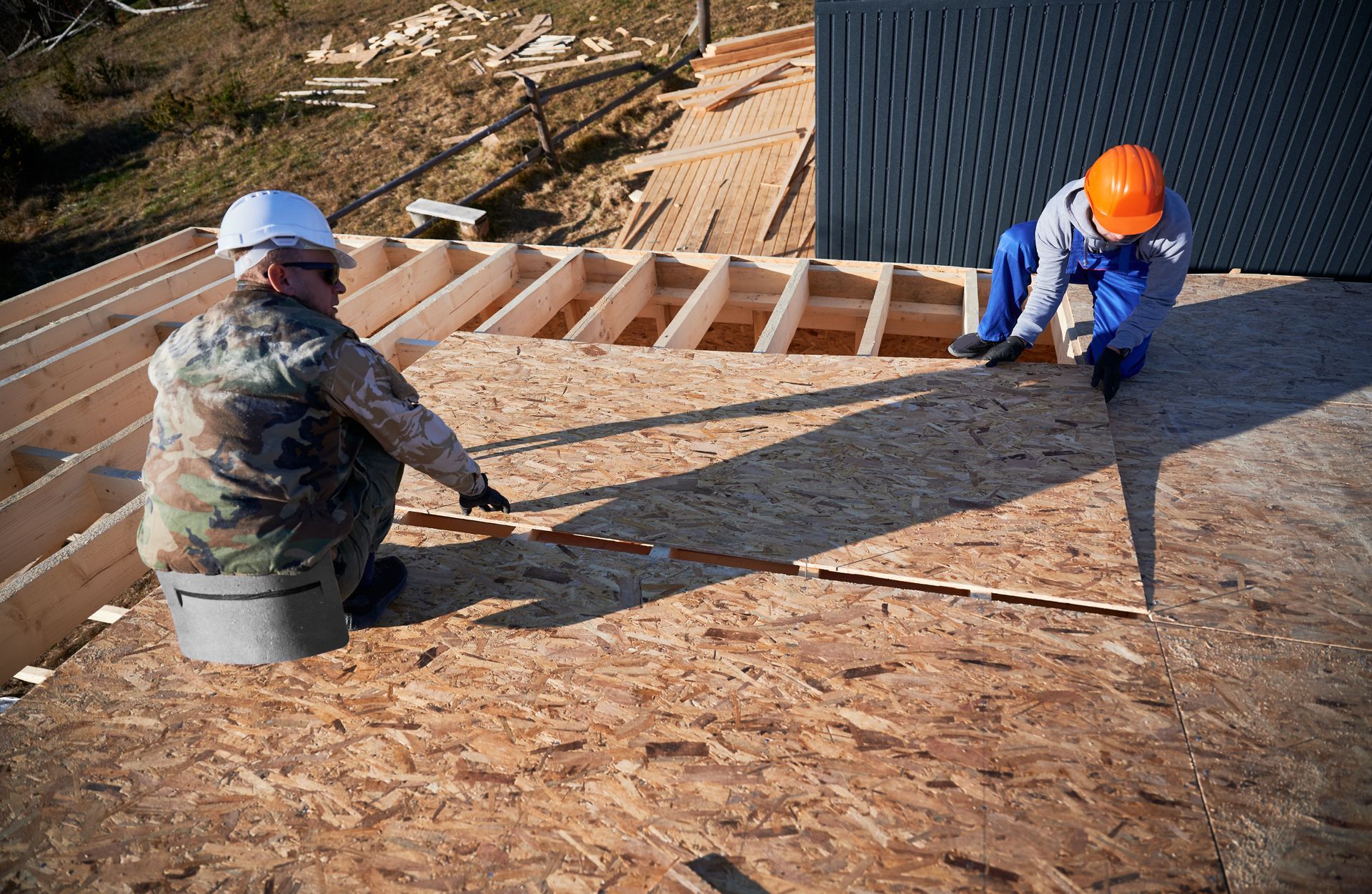 Two workers kneel on a plywood roof deck, installing panels beside exposed roof framing.