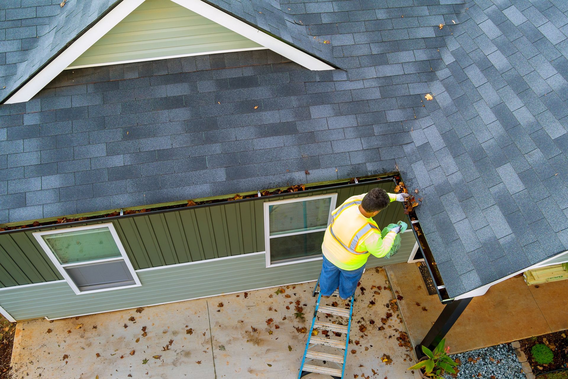 Worker on a ladder repairing roof shingles beside a house