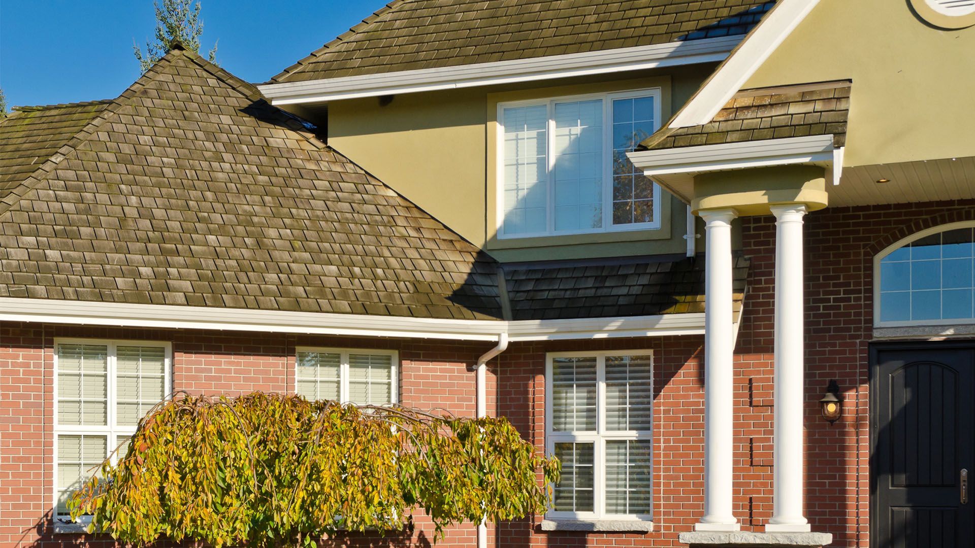 Brick house with multiple windows, columns, and a brown roof under a blue sky.