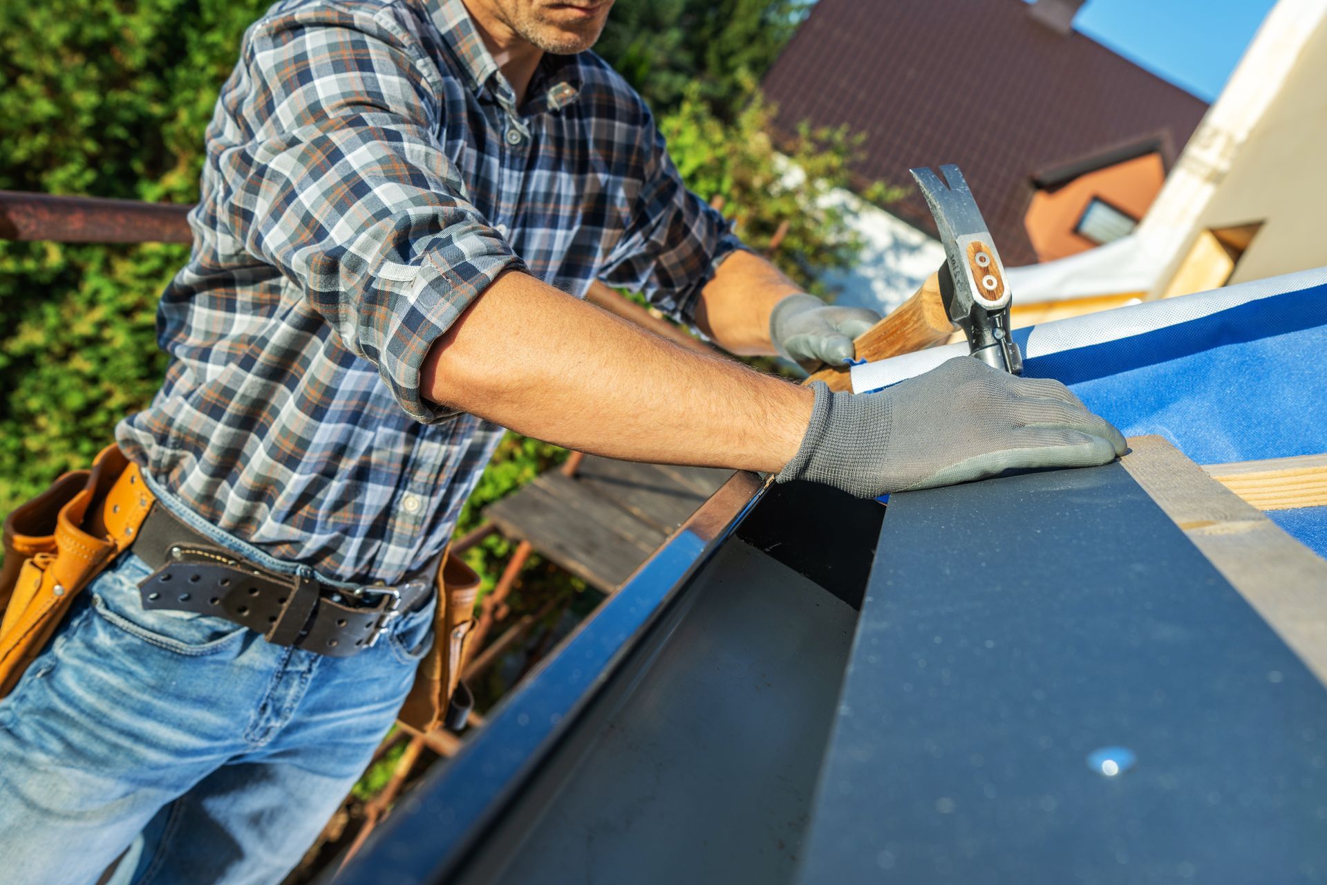 Roofer installing gutter with a hammer. Wearing gloves, plaid shirt, and jeans, working outdoors.