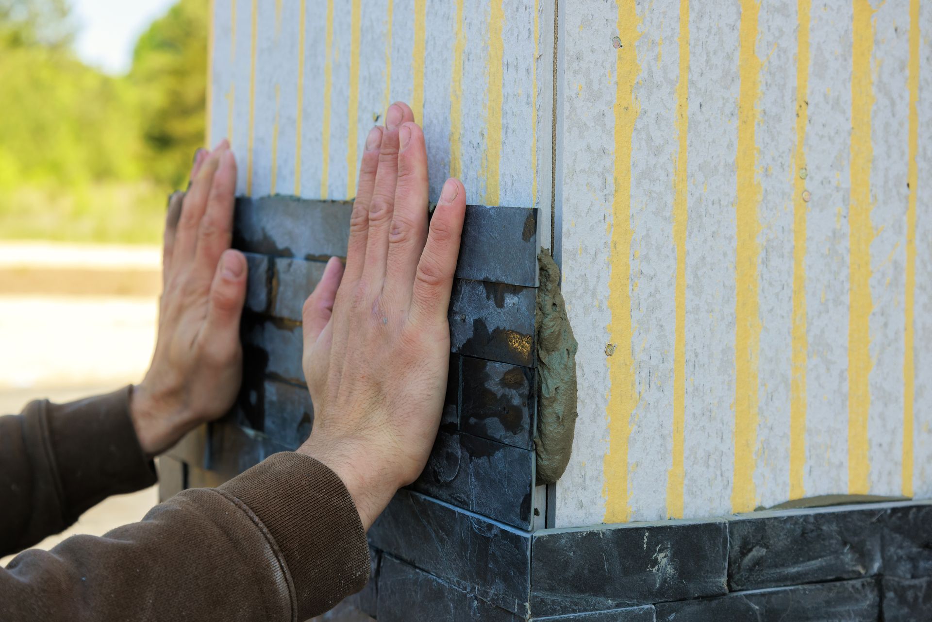 Hands pressing insulation panels onto an exterior wall with black waterproofing strip and yellow-striped wrap