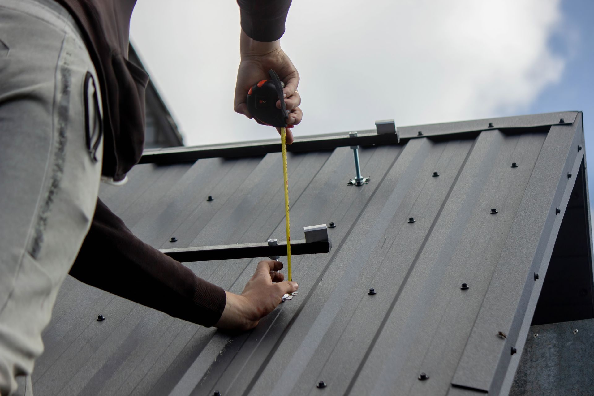 Two workers measuring a gray metal roof with a tape measure