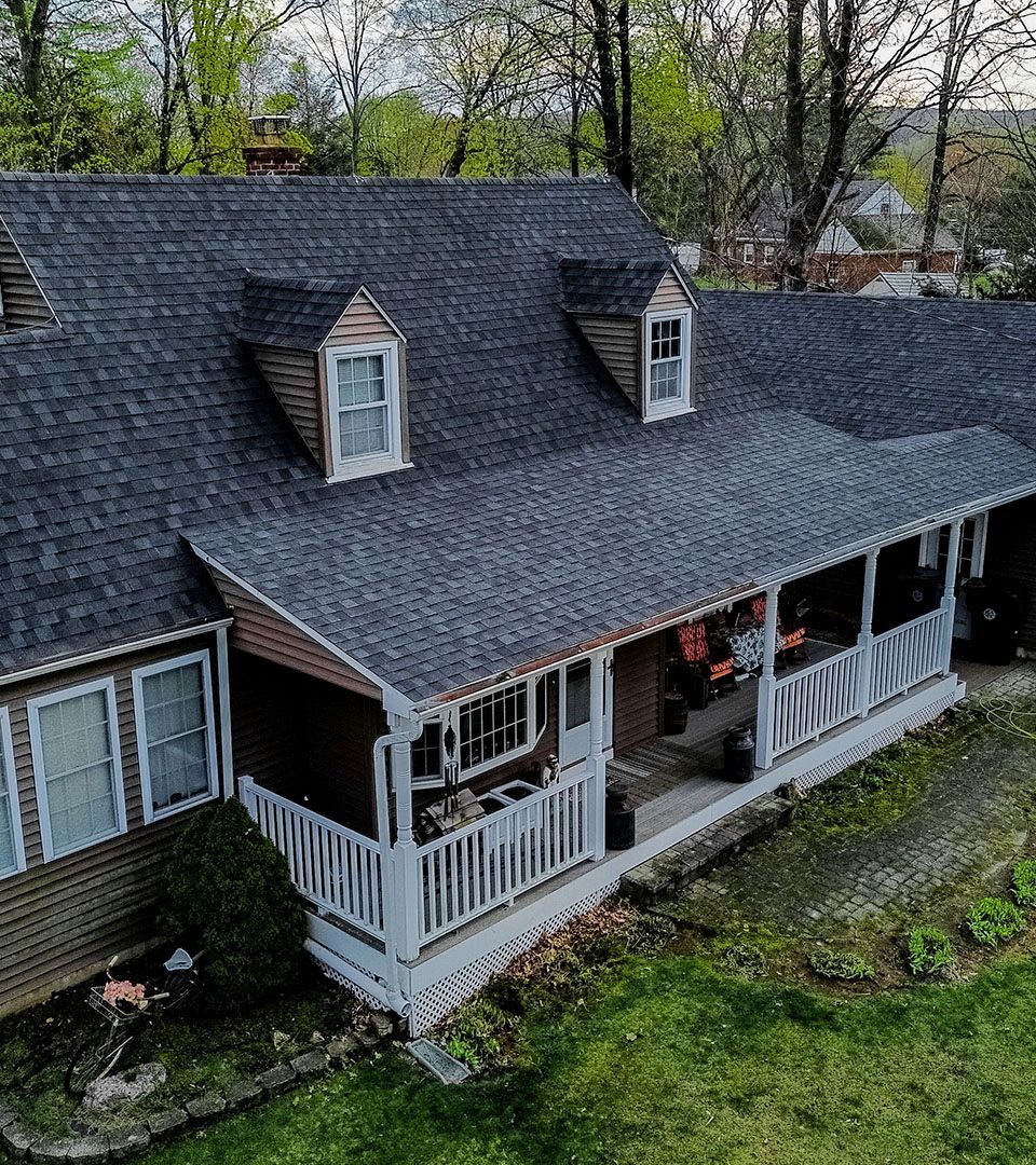 A brown house with a porch and dark gray roof, two dormers, and a white railing. Green grass surrounds.