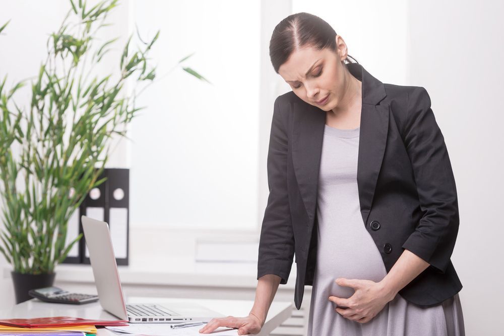 A pregnant woman is standing in front of a laptop computer holding her belly.